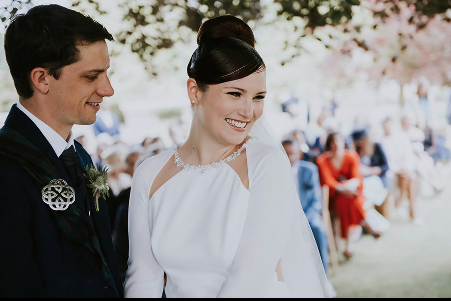 Bride smiles as groom looks at her at the altar, makeup by Jennifer Louise Studio.